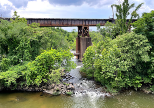 A rusted railway bridge spans a river surrounded by lush green trees and rocky banks under a cloudy sky.