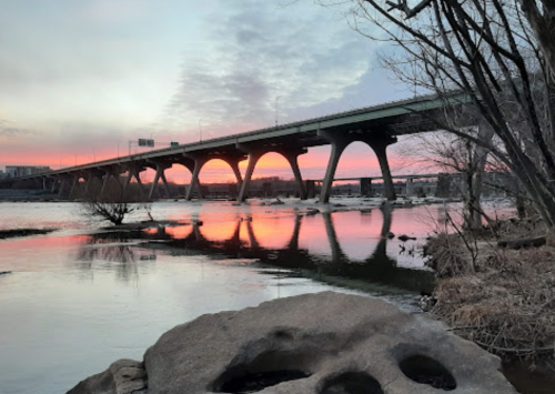 A bridge spans a river at sunset, reflecting vibrant colors in the water, with trees and rocks in the foreground.