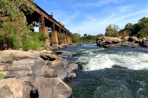 A river flows over rocks beneath a train bridge, surrounded by greenery and a clear blue sky.