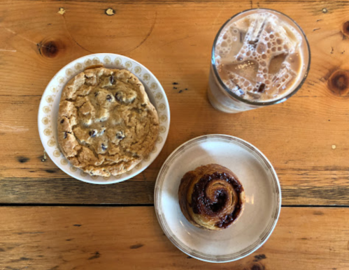 A plate with a chocolate chip cookie, a cinnamon roll, and a glass of iced coffee on a wooden table.