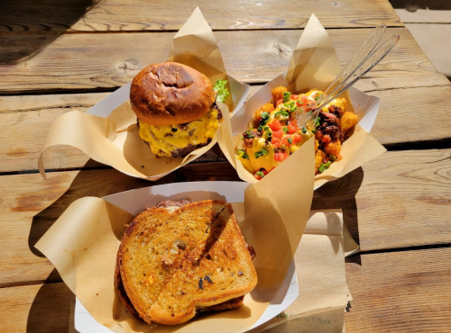 A wooden table with three food items: a burger, a sandwich, and a colorful salad with beans and vegetables.