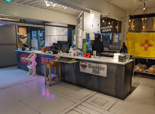 A food stall interior with a counter, menus on the wall, and a person preparing food in a vibrant, casual setting.