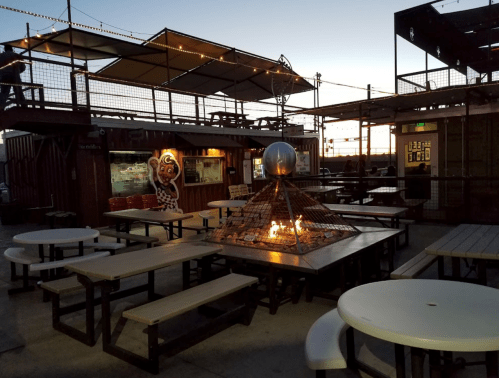 Outdoor seating area with picnic tables, a fire pit, and a colorful mural at sunset.
