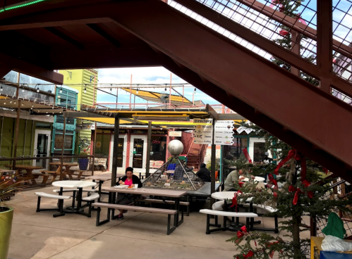 A vibrant outdoor seating area with colorful buildings, tables, and festive decorations, under a cloudy sky.