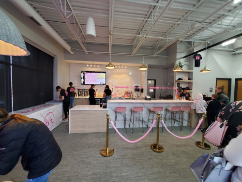 A modern café interior with a counter, menu screens, and customers in line. Pink decor and seating are visible.