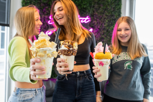 Three young women hold colorful milkshakes topped with various treats, smiling in front of a neon sign.
