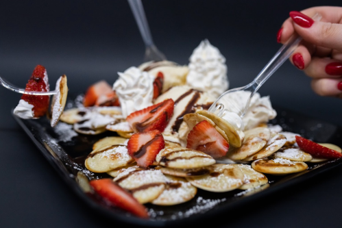A plate of mini pancakes topped with strawberries, whipped cream, and powdered sugar, with two forks digging in.