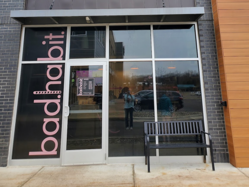 Exterior of a storefront with "bad.habit" signage, featuring a bench and large windows reflecting the surroundings.