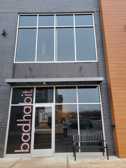 Exterior of a modern building with large windows and a sign reading "bad habit." A bench is visible in front.
