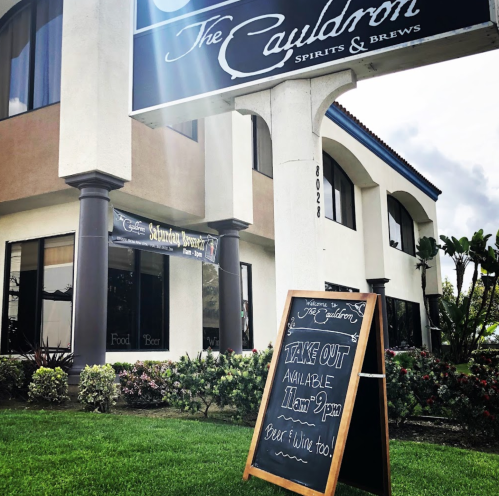 A sign outside "The Cauldron" restaurant announces takeout hours and offers food, beer, and wine.