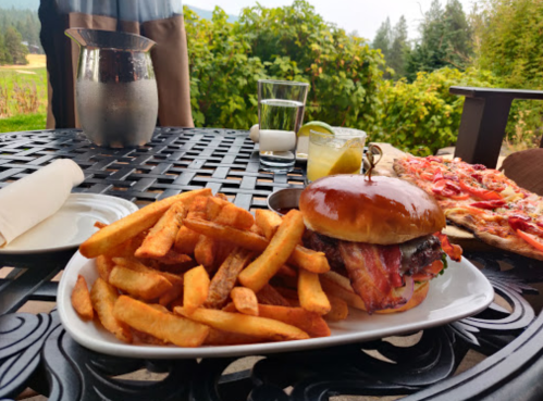 A plate with a burger topped with bacon, golden fries, and a side of drinks on a table outdoors.