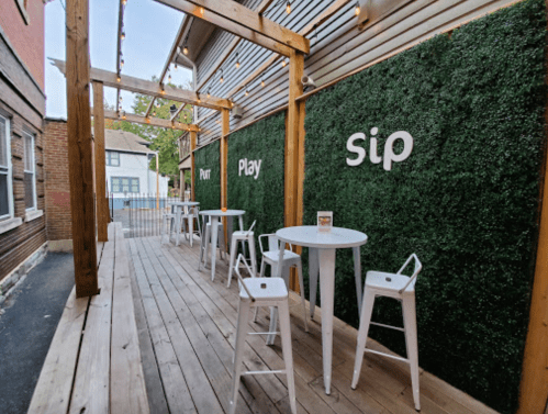 Outdoor seating area with wooden deck, green wall, and white tables and chairs, featuring the words "Pour," "Play," and "Sip."