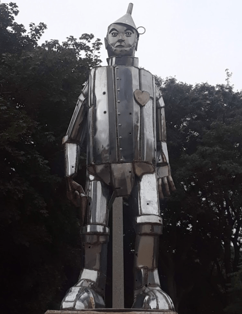 A large metallic statue of a humanoid figure with a heart, resembling a classic tin man, set against a tree backdrop.