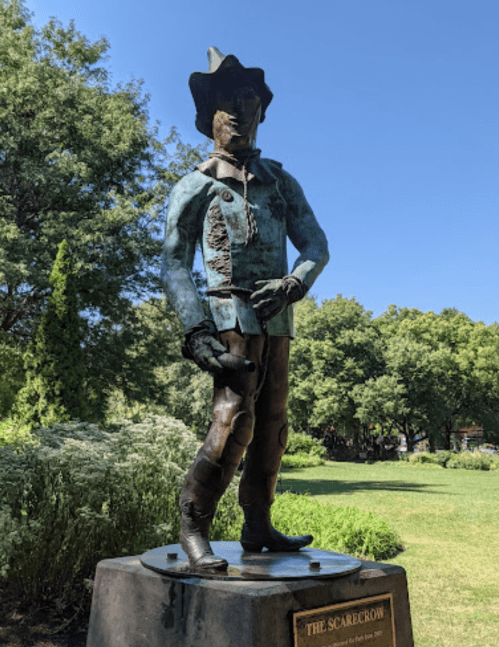 A bronze statue of a scarecrow in a cowboy hat, standing in a park surrounded by greenery.