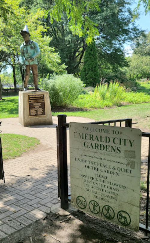 A statue stands in a park with a sign welcoming visitors to Emerald City Gardens, reminding them to respect the area.