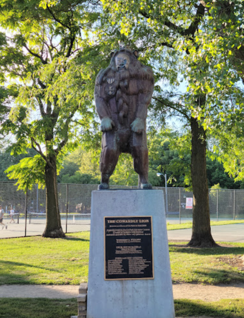 A bronze statue of a muscular figure standing on a pedestal, surrounded by trees and a park setting.
