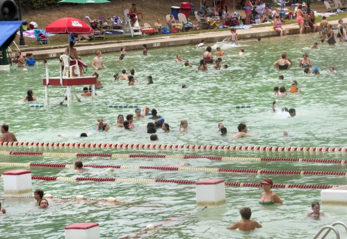 A crowded outdoor swimming pool with people swimming, lounging, and a lifeguard on duty under an umbrella.