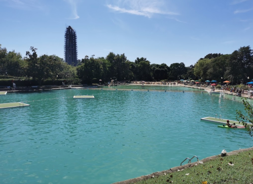 A serene blue lake surrounded by trees, with people enjoying the water and a tall building in the background.