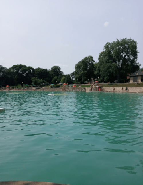 A serene lake with turquoise water, surrounded by trees and a sandy shore, with people enjoying the area in the background.