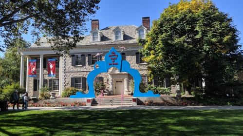 A large stone house with a blue archway entrance, surrounded by greenery and colorful banners.