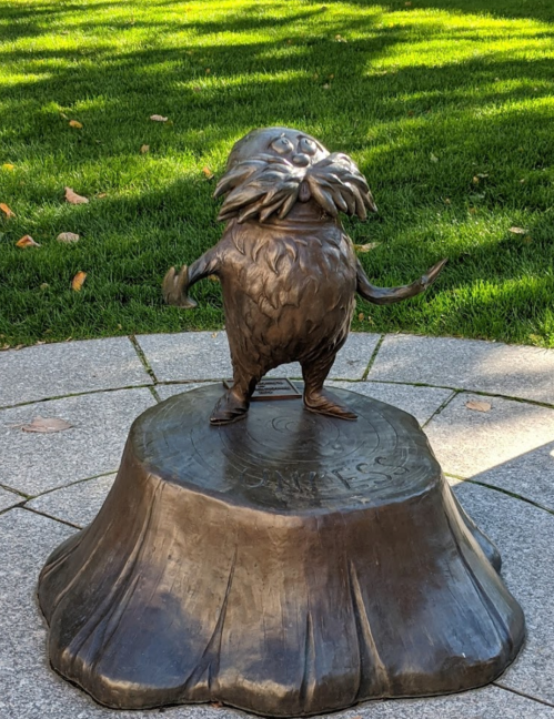 A bronze statue of a whimsical creature standing on a tree stump, surrounded by green grass.