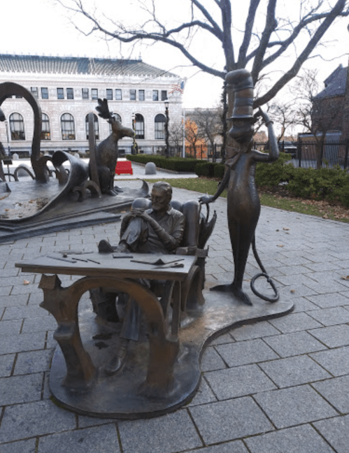 A whimsical bronze sculpture featuring a seated man at a desk and a tall, hat-wearing character beside him in a park.