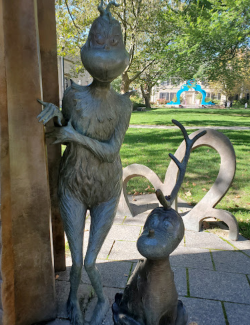Bronze sculptures of the Grinch and a small reindeer in a park setting with trees and a fountain in the background.