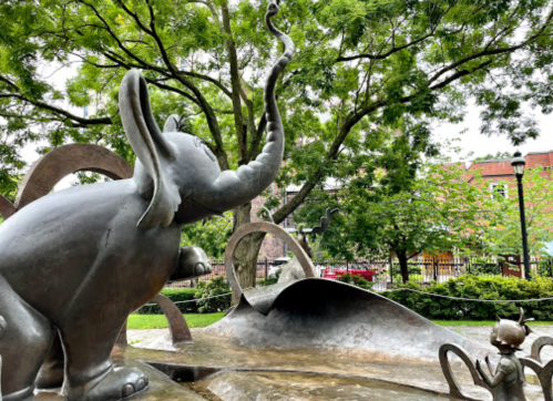 A bronze sculpture of an elephant and a child, surrounded by trees and greenery in a park setting.
