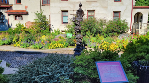 A garden scene featuring a tall sculpture surrounded by colorful flowers and greenery, with a sign nearby.
