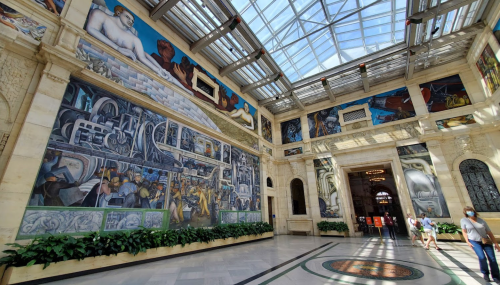 A spacious gallery featuring large murals, bright skylights, and lush greenery along the walls. Visitors explore the area.