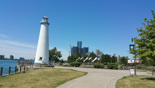 A white lighthouse beside a waterfront path, with a city skyline in the background under a clear blue sky.