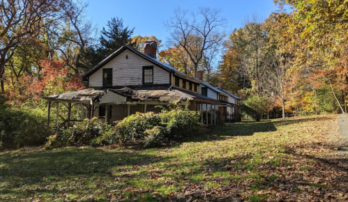 A weathered, two-story house surrounded by autumn foliage and fallen leaves under a clear blue sky.