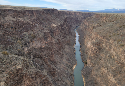 A winding river flows through a deep canyon surrounded by rocky cliffs and sparse vegetation under a cloudy sky.