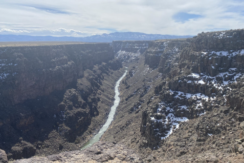 A deep canyon with a winding river at the bottom, surrounded by rocky cliffs and snow-capped mountains in the distance.