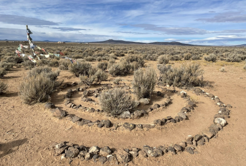 A spiral stone formation in a desert landscape, surrounded by sparse vegetation and distant mountains under a cloudy sky.