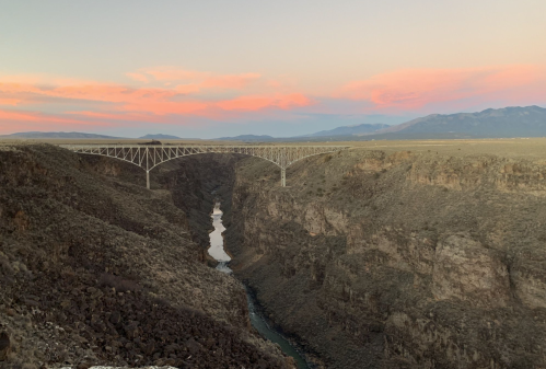 A scenic view of a bridge spanning a deep canyon at sunset, with mountains in the background and a river below.