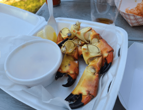 A takeout container with crab claws, lemon wedges, and a small cup of dipping sauce on a wooden table.