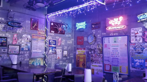 A vibrant bar interior with neon signs, colorful posters, and a lively atmosphere, featuring a table and chairs.