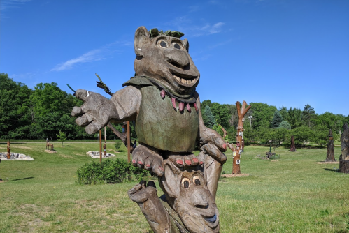 A large wooden sculpture of a whimsical creature with a playful expression, set in a grassy park with trees in the background.