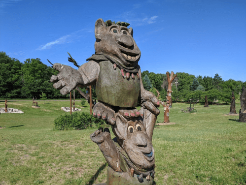 Two whimsical wooden sculptures of trolls, one perched on the other's shoulders, set in a grassy park.