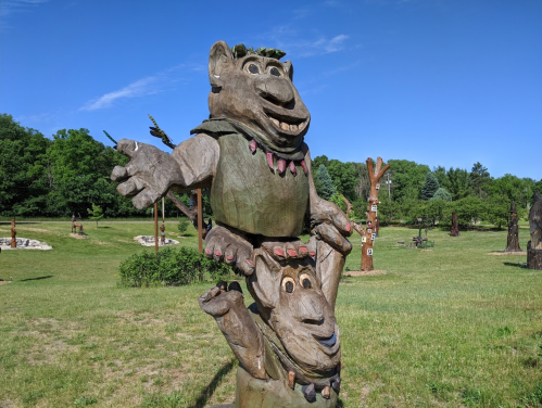 Two whimsical wooden sculptures of trolls, one perched on the other's shoulders, set in a grassy park.