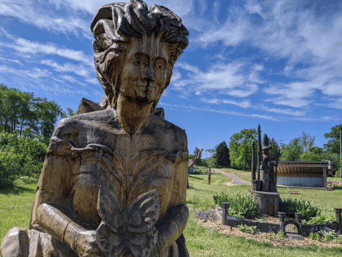 Wooden sculpture of a woman holding a butterfly, set against a blue sky and green landscape.