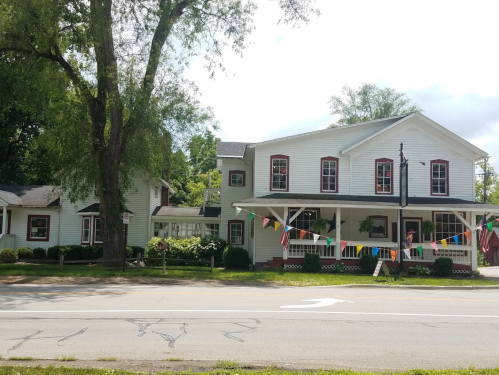 A white two-story building with red accents, surrounded by greenery and colorful flags, along a quiet road.