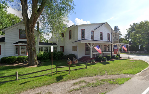 A charming white building with a porch, surrounded by greenery and American flags, set against a clear blue sky.