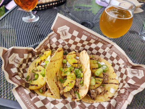 Three grilled tacos filled with meat, topped with green onions and pineapple, served on a checkered paper.