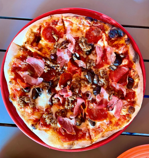 A round pizza topped with pepperoni, sausage, ham, olives, and melted cheese on a red plate.