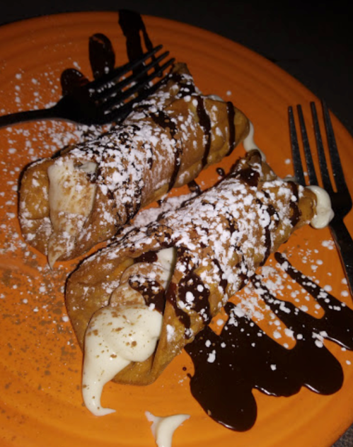 Two dessert tacos filled with cream, drizzled with chocolate sauce, and sprinkled with powdered sugar on an orange plate.