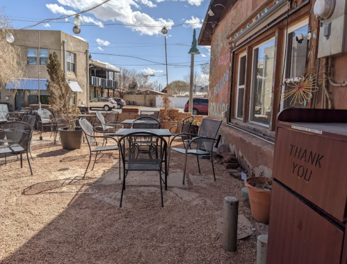 Outdoor seating area with metal chairs and tables on gravel, surrounded by buildings and a blue sky with clouds.