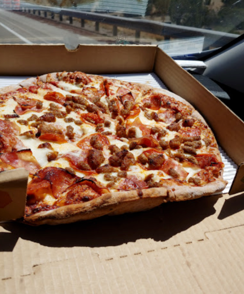 A large pizza with pepperoni and sausage in an open pizza box, sitting on a car seat with a road in the background.