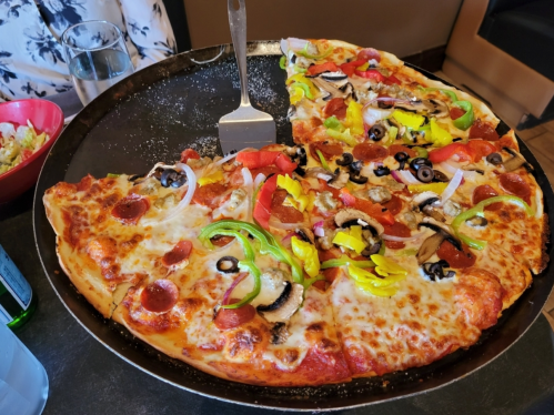 A large pizza with various toppings, including pepperoni, vegetables, and olives, served on a round tray.